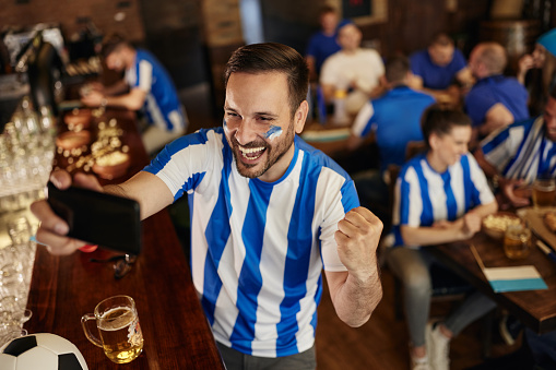 Happy soccer fan taking a selfie with smart phone after watching a successful game in a bar.