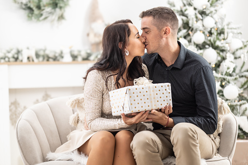 Woman and man kiss in front of the Christmas tree holding a Xmas present together.