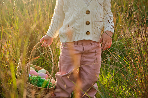 Crop unrecognizable girl in white sweater carrying colorful Easter eggs in wicker basket while standing on grassy lawn in sunny countryside