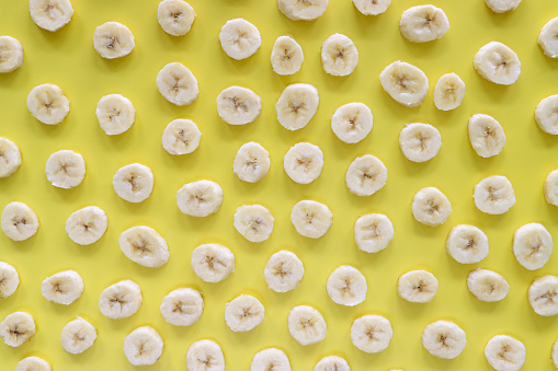 Stock photo showing close-up, elevated view of layer of banana fruit slices on a yellow background, healthy eating poster wallpaper background design.