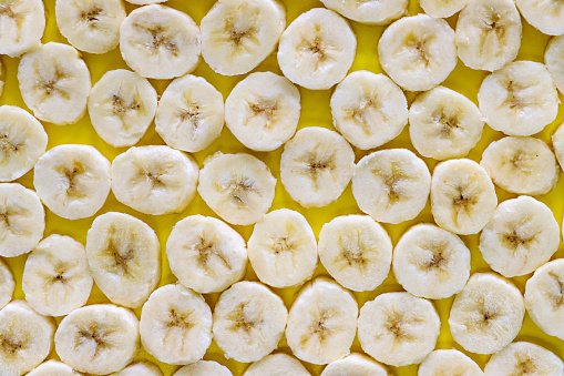 Stock photo showing close-up, elevated view of layer of banana fruit slices on a yellow background, healthy eating poster wallpaper background design.