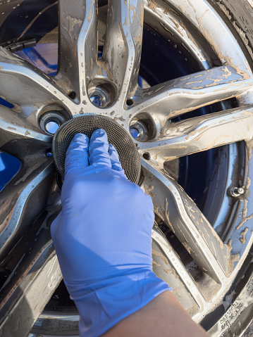 Hand wearing a blue glove scrubbing a dirty alloy wheel with silicone sponge. First person point of view.