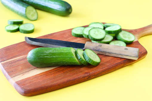 Stock photo showing close-up view of healthy eating image of a wooden chopping board covered in cucumber slices on yellow background. Pieces of cucumber (Cucumis sativus) displaying green skin and white flesh with white seeds.