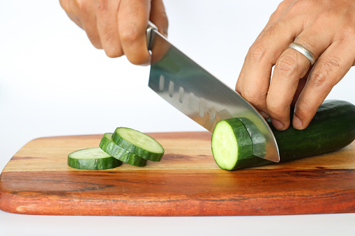 Stock photo showing close-up view of healthy eating image of a wooden chopping board covered in cucumber slices on white background. Pieces of cucumber (Cucumis sativus) displaying green skin and white flesh with white seeds.