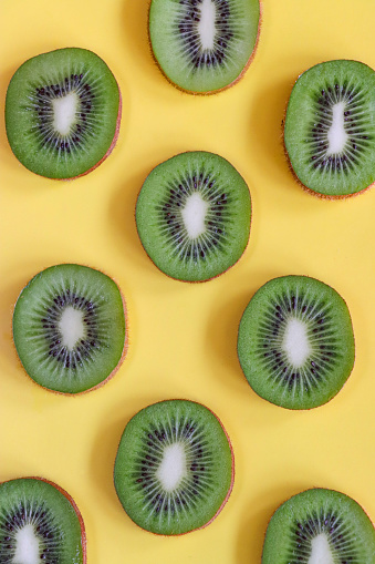 Stock photo showing an elevated view of a layer of kiwi fruit slices on a yellow background. Pieces of Chinese gooseberry displaying fuzzy brown skin and bright green flesh with ring of black seeds, healthy eating poster wallpaper background design.