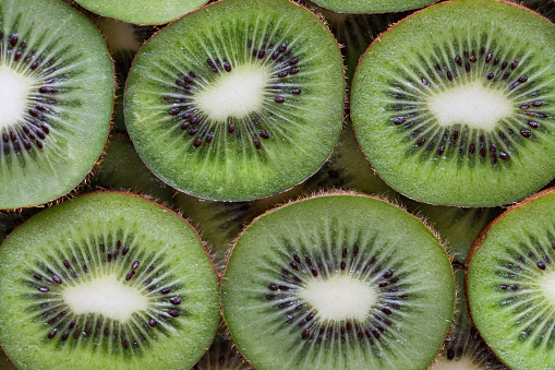 Stock photo showing close-up, elevated view of layers of kiwi fruit slices. Overlapping pieces of Chinese gooseberry displaying fuzzy brown skin and bright green flesh with ring of black seeds, healthy eating poster wallpaper background design.