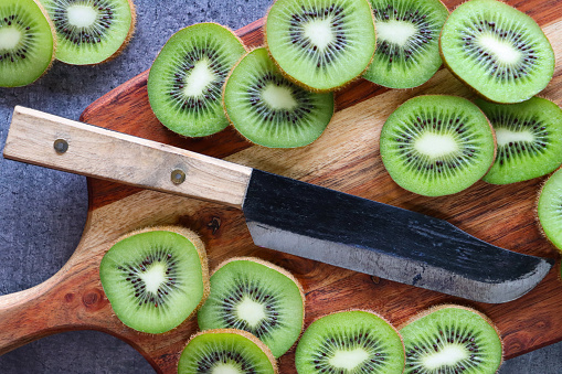 Stock photo showing a close-up view of healthy eating image of a wooden chopping board covered in slices of kiwi fruit on a mottled grey background. Pieces of Chinese gooseberry displaying fuzzy brown skin and bright green flesh with ring of black seeds.