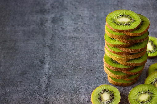 Stock photo showing a close-up view of healthy eating image of a pile of kiwi fruit slices displaying fuzzy brown skin and bright green flesh with ring of black seeds, on mottled, grey background.