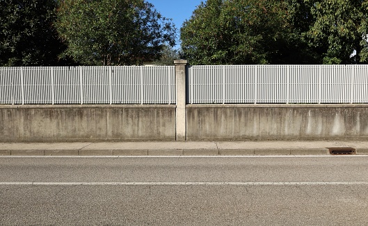 Fence made of concrete wall with white railing on top. Decorative column in the middle. Trees on behind. Cement tiled sidewalk and street in front. Background for copy space.
