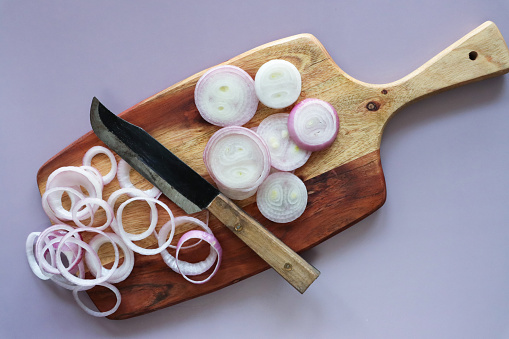 Stock photo showing a close-up view of healthy eating image of a wooden chopping board covered in red onion in various states of being cut up. Rings and sliced pieces of vegetables displaying purple skin and white flesh.