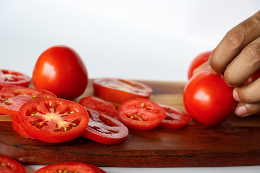 Stock photo showing a close-up view of healthy eating image of a wooden chopping board covered in tomatoes in various states of being cut up. Whole, halved and sliced pieces of fruit displaying red skin, flesh and seeds.