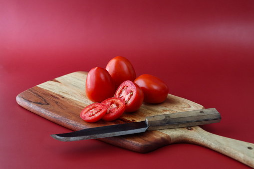 Stock photo showing a close-up view of healthy eating image of a wooden chopping board covered in tomatoes in various states of being cut up. Whole, halved and sliced pieces of fruit displaying red skin, flesh and seeds.