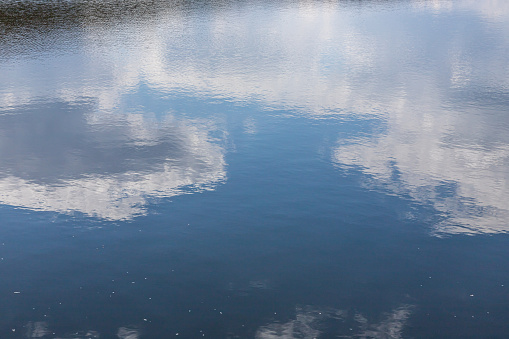 Calm water surface. Summer evening near a forest lake, the surface of the water is smooth, with small waves. The sky with clouds is reflected from the surface, coloring the water in different colors.