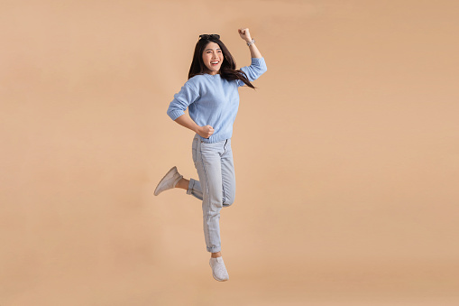Happy smiling Asian woman with jumping in the air lifting her hands up in blue sweater and jeans isolated on beige color background. Full body length in studio shot.