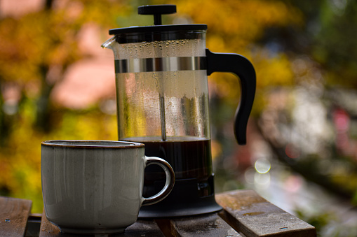 A cup of coffee and a french press coffee pot placed on top of a wooden table