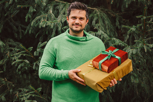 Stylish man in green sweate with Christmas gift on pine background