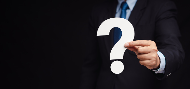 Question mark symbol. Businessman in a suit holding a white question mark paper while standing on a black background. Close-up photo. Space for text