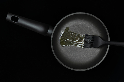 Black greasing silicone brush smears oil on frying pan. Concept for cooking, kitchenware advertising, Black Friday presentation. Top view, flat lay, black background.