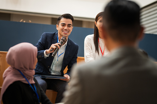 Shot of a multiracial group of businessperson having a seminar in a modern office. Young businessman gestures while asking a question during a Q&A portion of a business conference. - Business conference APAC