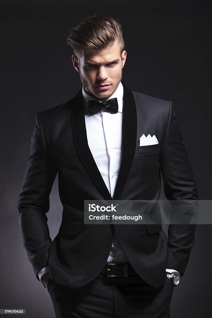 business man holds both hands in pockets elegant young fashion man in tuxedo holding both his hands in his pockets while looking at the camera with a frowned expression. on black background Adult Stock Photo