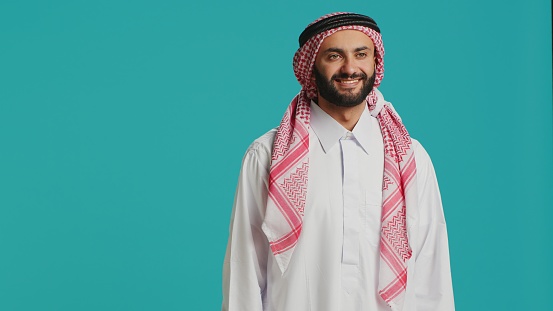 Middle eastern guy in traditional wear, beaming and standing proudly while having a checkered pattern headpiece and a white gown, illustrating islamic culture. Young delighted arab man.