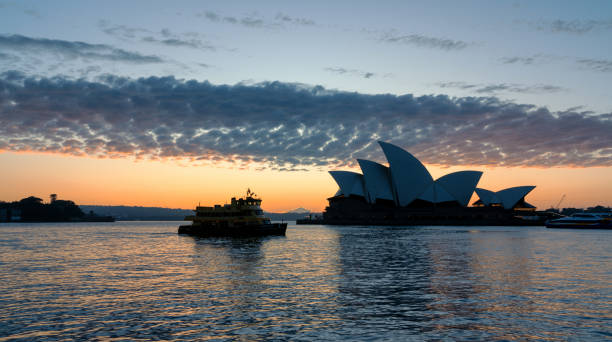 Sydney Australia - August 29, 2023: Early morning in Sydney, and the iconic Opera House and a glorious sunrise are reflected in the waters of Circular Quay. A silhouetted ferry can be seen steaming past the Opera House.