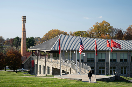 this color image is of a School Building or Business Building with American Flag. the building could also be a library or mall or church or university or gymnasium. the architecture is very modern. there is a massive sidewalk or walkway to the building. and the entrance is made of glass. the building is made of brick, glass and cement. there is also a lawn that is of green grass. and the lighting is natural sunlight. and the background is blue sky with white clouds. 