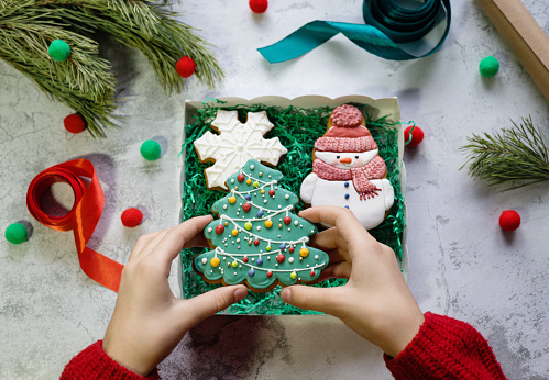 Girl's hands in red knitted sweater put gingerbread in the form of  Christmas tree into gift box with snowman and snowflake gingerbreads next to pine branch, ribbons and wrapping paper.