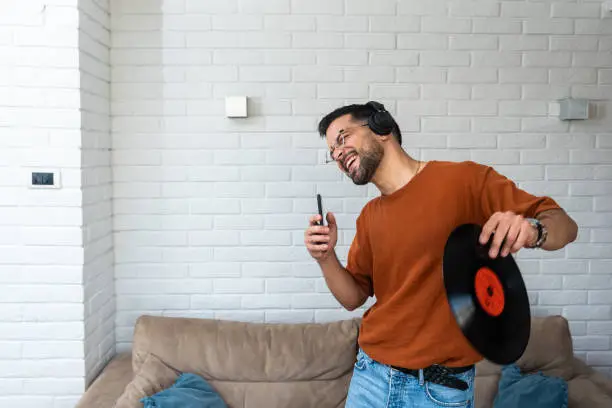 Photo of Young happy man singing and dancing at home listening music on wireless headset singing on his phone holding LP record in his hand. Hipster music lover digitized old record audio and enjoying