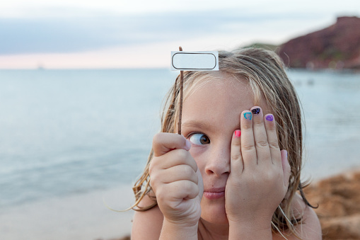 Discover pure joy in 'A Very Young Girl with Blond Hair on the Beach.' With a carefree spirit, the little one makes a funny face while holding a white label, creating a whimsical moment against the backdrop of the sunlit beach. A delightful snapshot of innocence and seaside playfulness.