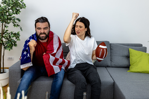A young couple is enjoying beer and popcorn while watching a rugby game and feeling enthusiastic about holding the US flag.
