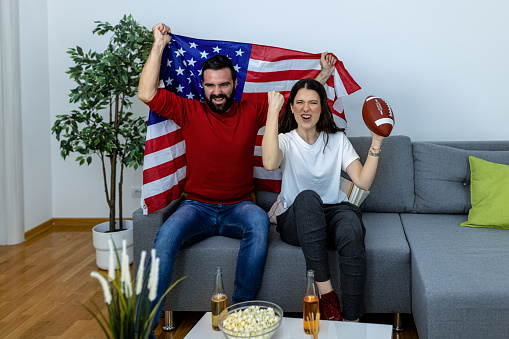 A couple of young friends are sitting in the living room, drinking beer and supporting their favorite football team with the national flag.