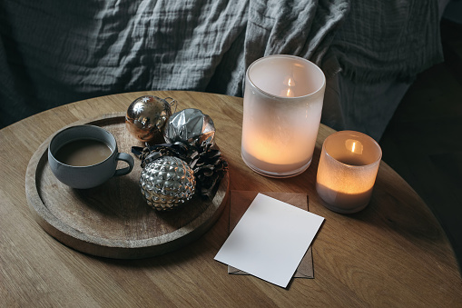 Moody cozy winter home, interior still life. Cup of coffee, vintage Christmas ornaments, balls on round wooden table. Blurred background. Candles in colorful glass candleholders, blurred background.