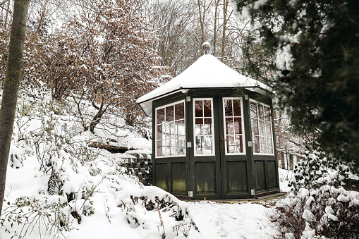 Old dark green gazebo in outdoor, park. Little wooden garden shelter, cabin covered with snow. Beautiful winter garden, landscape. Season in nature. Snowy trees, ground, blurred foreground, no people.