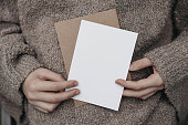 Winter, autumn cozy stationery still life. Child, female hands in beige pullower holding blank greeting card, craft envelope. Invitation mockup on knitted wool sweater. Blurred background.