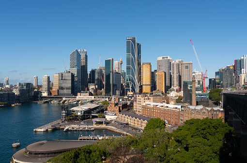 Circular quay of Sydney city on shore of Sydney Harbour with tall high-rise office and business towers over wharves, ferries and train station.