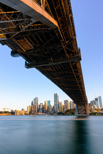 The spectacular cityscape of Sydney, Australia, as seen from beneath the iconic Harbour Bridge. Across the water we see the iconic Sydney skyline featuring the towering skyscrapers of the CBD.