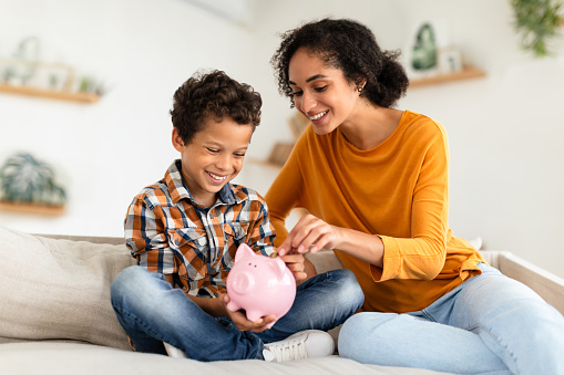 Economy, Kid Personal Savings. Happy Mom And Little Son Putting Coin In Piggybank Sitting In Modern Living Room At Home. Financial Investments Safety, Great Bank Offer Concept. Selective Focus