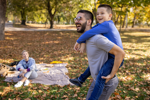 A mid adult Caucasian man carrying his son on his back while on a picnic with his family in a public park