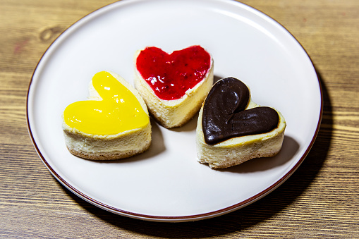 Three color cheesecakes heart shape lemon chocolate and raspberry on white plate on wooden background closeup top view