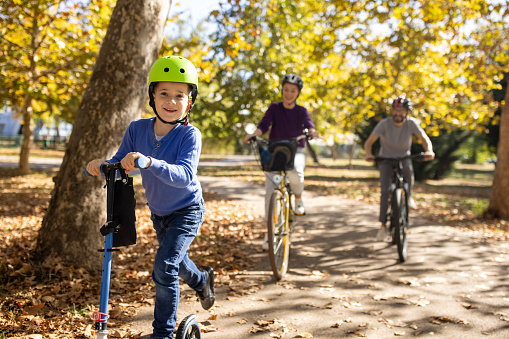 A young Caucasian boy riding a push scooter while his mother and father ride bikes with him in a public park