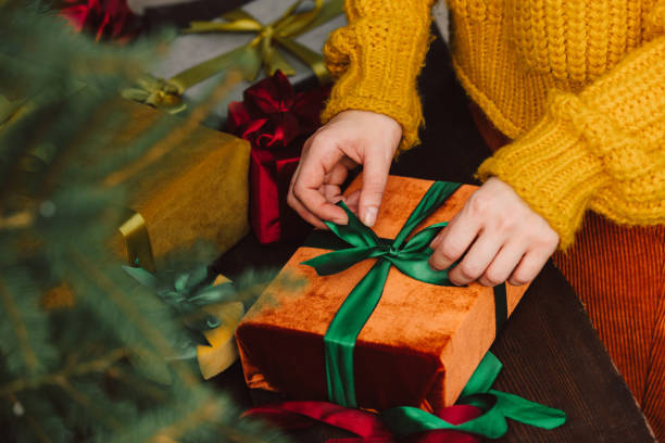 Woman in yellow sweater wrapping fabric eco friendly Christmas gift boxes on wooden table next to Christmas tree stock photo