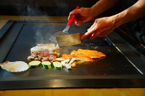 chef's hands with spatula over teppanyaki. cooking vegetables meat and seafood on hot hibachi grill table. Traditional Japanese Cuisine. Teppan show