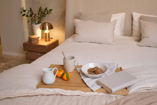 Mug of coffee mandarins cookies and book placed on wooden food tray on bed in cozy bedroom