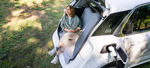 Woman with smartphone sits in an electric car's trunk.