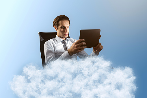 Young caucasian businessman using tablet on cloud. Blue sky background. Dream big, business and strategy, forethought and vision concept