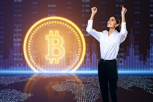 Cheerful businesswoman with raised hands standing near glowing bitcoin symbol on digital background. Cryptocurrency concept