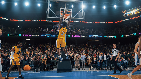 African American National Basketball Superstar Player Scoring a Powerful Slam Dunk Goal with Both Hands In Front Of Cheering Audience Of Fans. Cinematic Sports News Shot with Back View Action