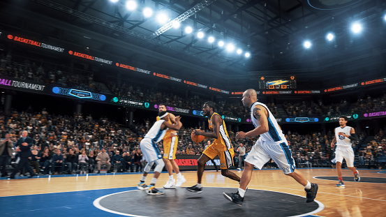 Low angle view of a professional basketball game. A player is in mid air holding ball about to score a slam dunk, but the player from the opposite team is ready to block him.  A  game is in a indoor floodlit basketball arena. All players are wearing generic unbranded basketball uniform.