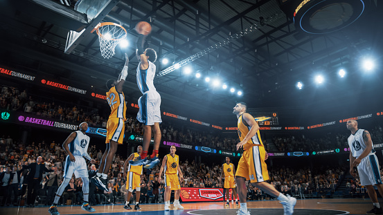 Image from a low angle viewpoint of a basketball player going to shoot a ball in a hoop  basketball in front of rival players. The action takes place in generic indoor floodlit basketball arena full of spectators. All players are wearing generic unbranded basketball uniform.